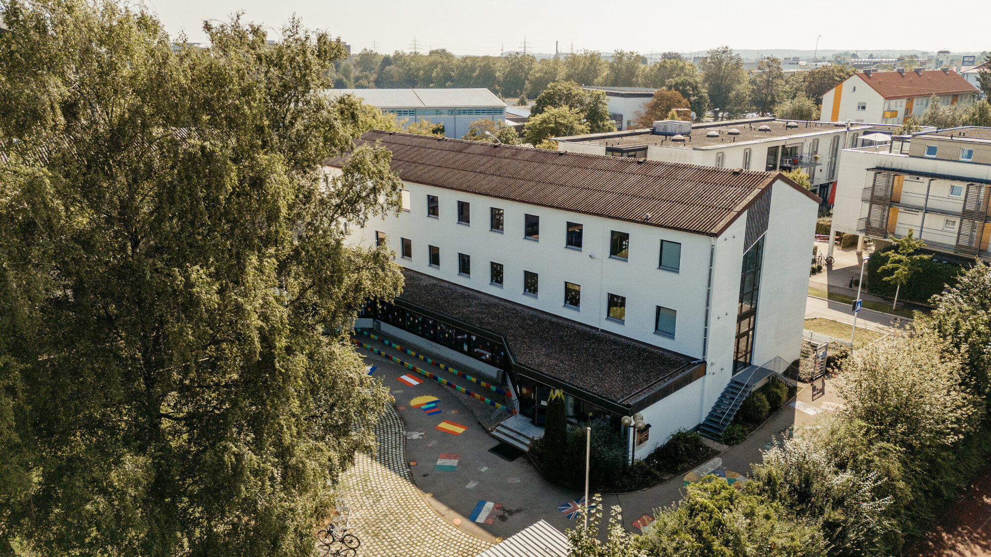 Luftaufnahme des weißen Schulgebäudes der St.-Michael-Grundschule in Neu-Ulm mit buntem Pausenhof
