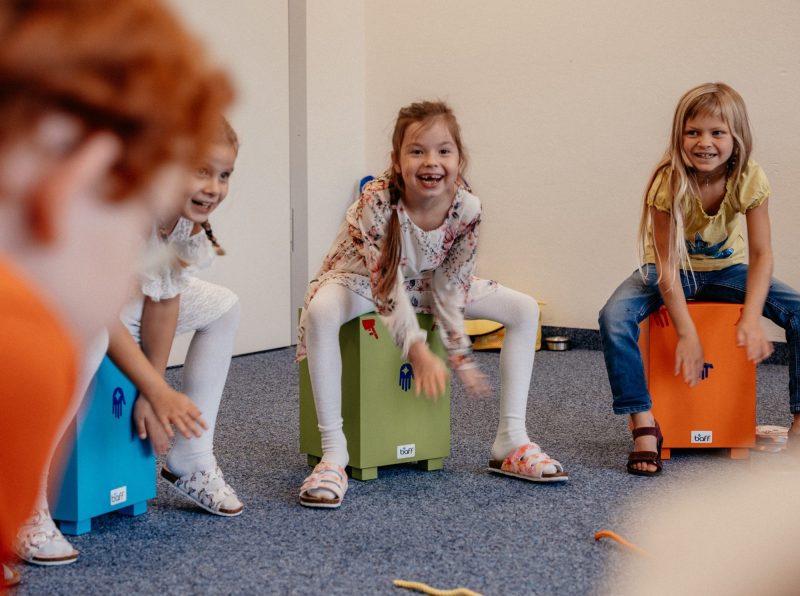 Schülerinnen spielen lachend auf bunten Cajons im Musikunterricht der St.-Michael-Grundschule Neu-Ulm